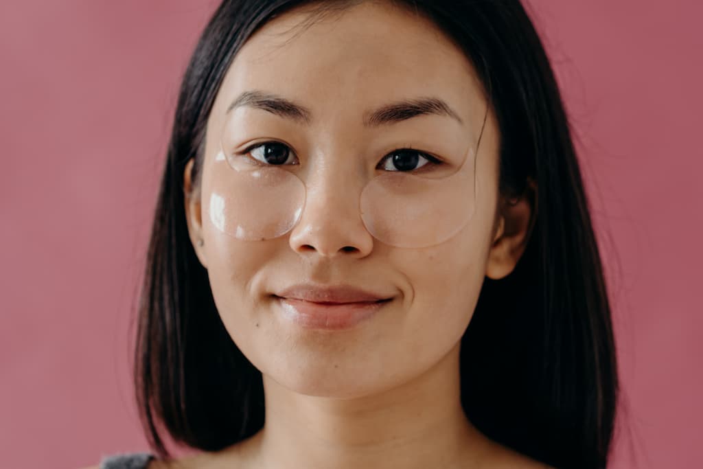 Japanese Skincare 1 A young asian women is in front of a dusty pink bacjground as she gazes striaght at you with a relaxed smile and eye masks laying just underneath her eyes