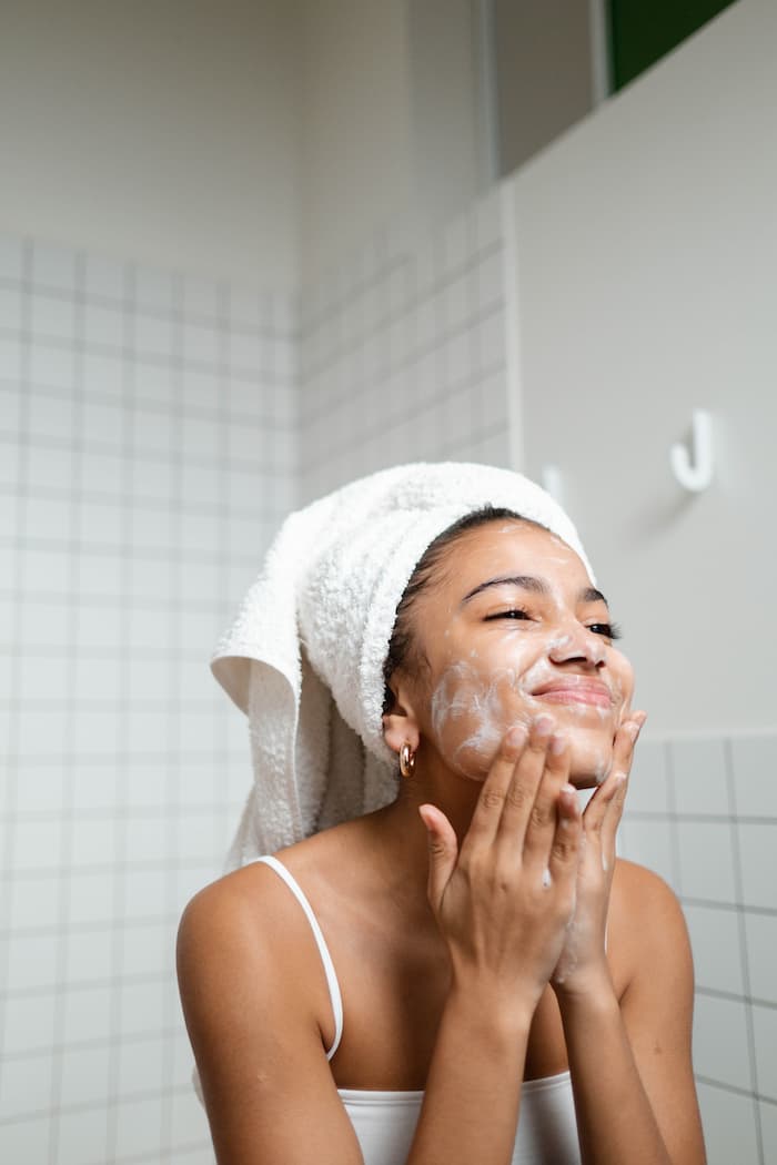 Japanese Skincare 1 A young black women is in her bathroom wearing a white tank top with her hair wrapped up in a white towel as she washes her face and is smiling at herself in the mirror