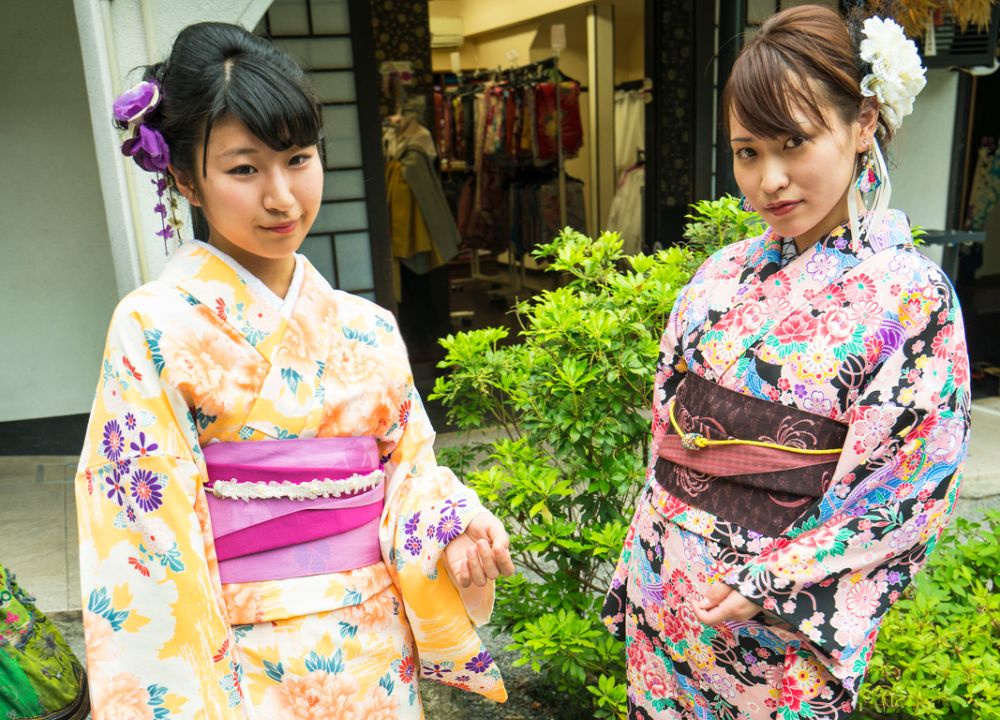 Kimono Rental 2 Two Japanese girls standing on the street wearing a yellow kimono with a pink obi (belt) and a pink kimono with a brown obi