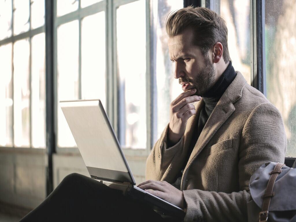 A man in a suit being confused by the usage of Japanese proverb, while sitting by the window with his laptop.