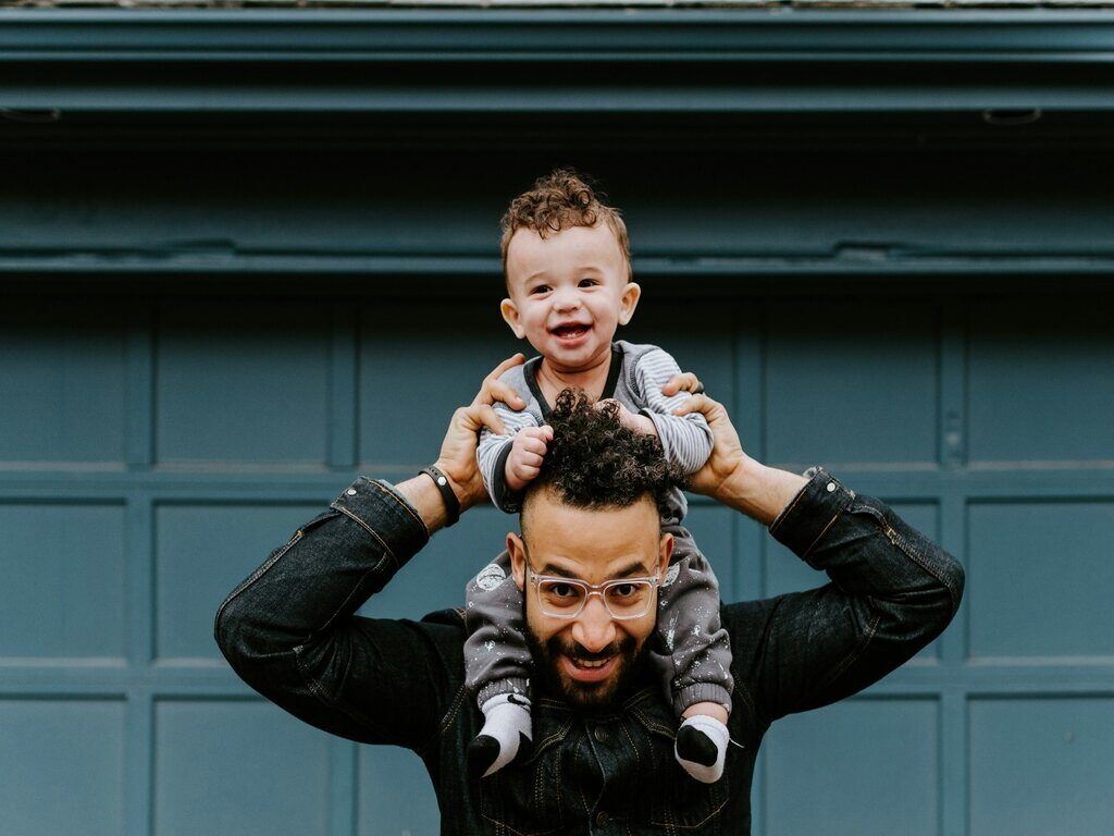 A smiling baby riding on his father's shoulders. Both are facing towards the camera. It is a visual image of the kotowaza "Kaeru no ko ha kaeru." or "Like father, like son."