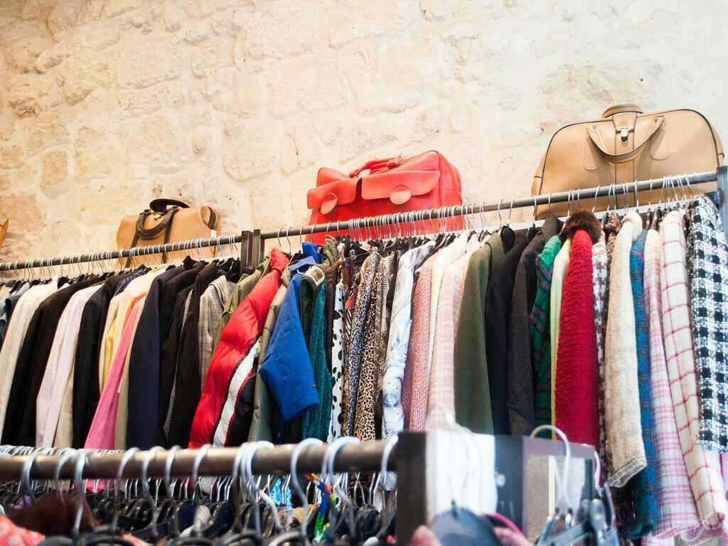 Various clothing hanging on a clothing rack in a Japanese thrift store. There are also three bacg placed on the rack.