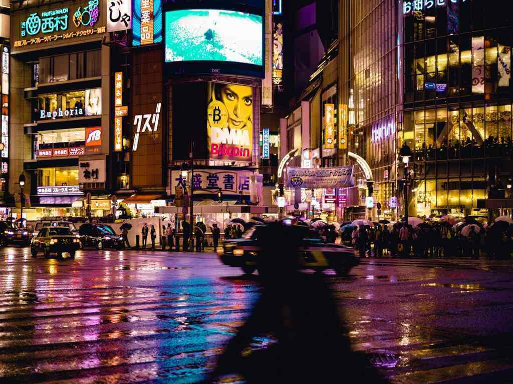 Shibuya scramble crossing at night. The road is wet due to the rain and reflects lights from the advertising signs.