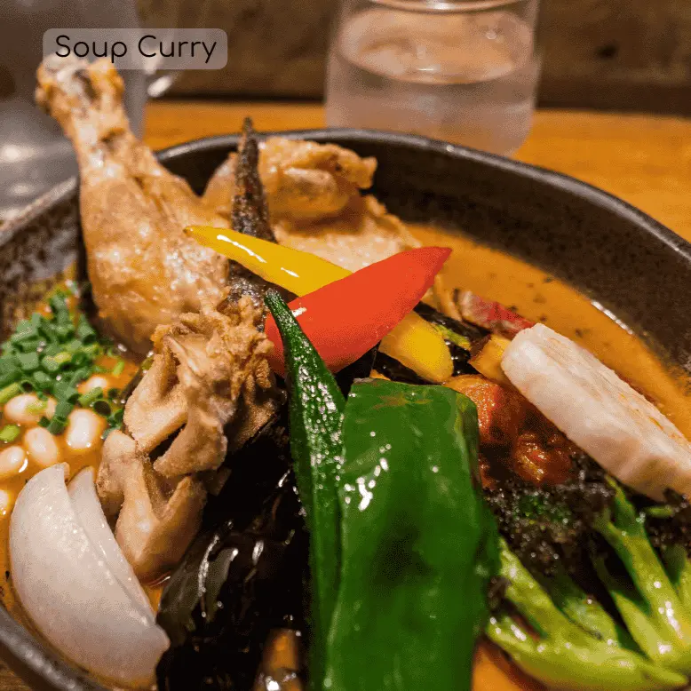 An image of soup curry in a bowl with chicken and a variety of vegetables such as green pepper, red pepper, broccoli, and onions.